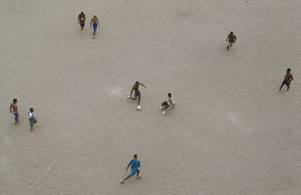 looking down on a football match played on dusty beach with man just about to kick a ball,