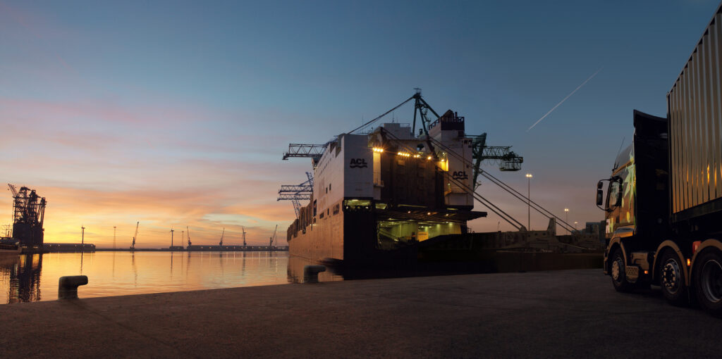 docks, with sunset over sea with cranes . Drive on Boat in foreground, with RH truck ready to drive on.