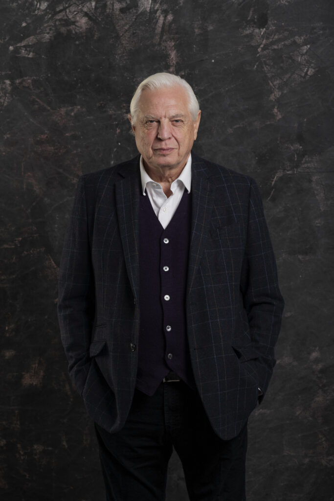 John Simpson standing in front of a black textured background looking direct to camera. Photograph by Gullachsen