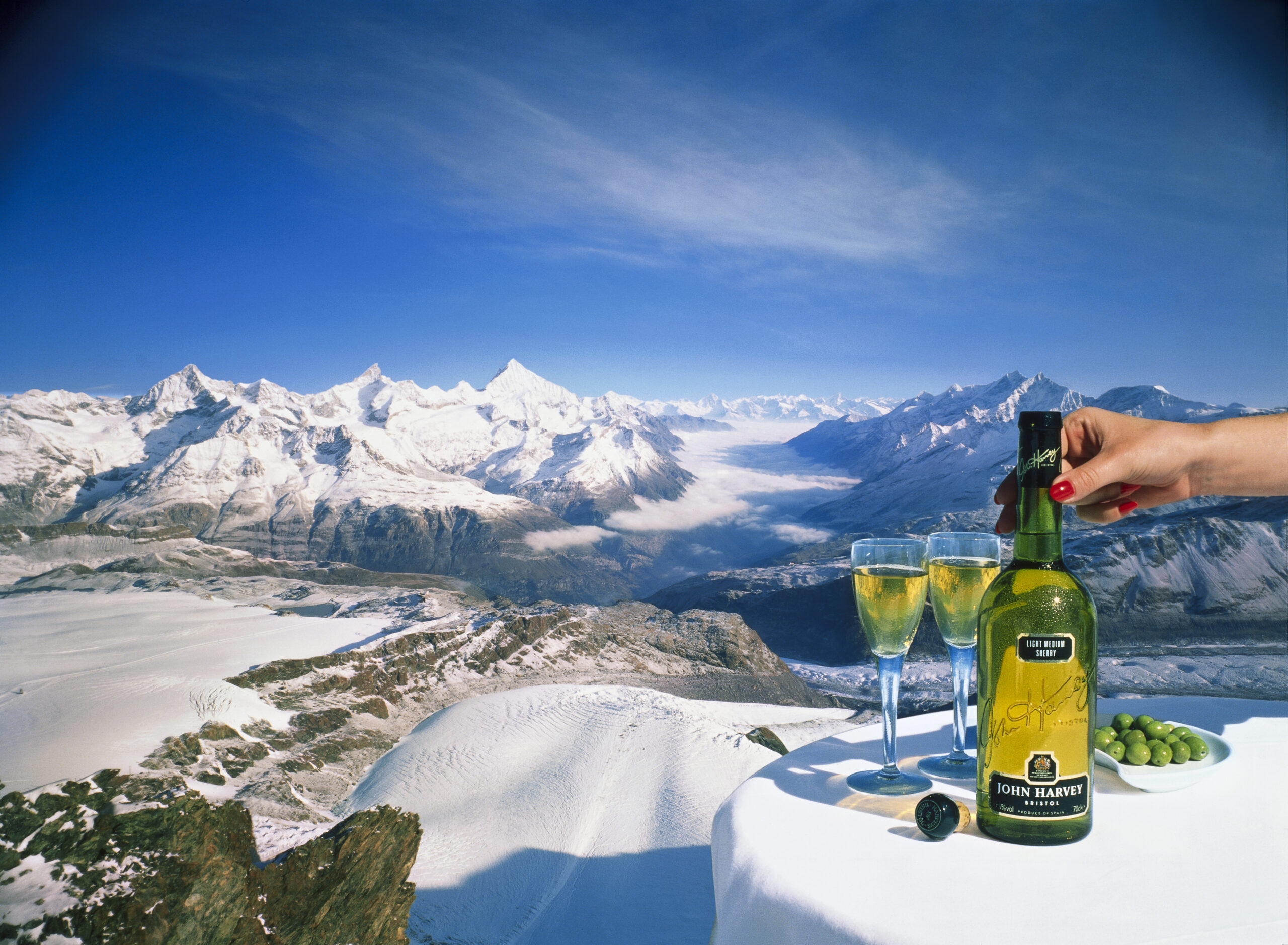 View from Mountain to looking down to valley with clouds and snow capped mountains around, Lady holding Sherry Bottle with glasses and olives on white tablecloth on table