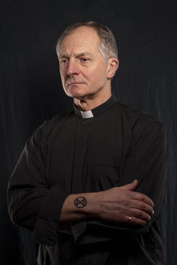 man in black with a clergy dog collar and tatoo on hand with symbol X