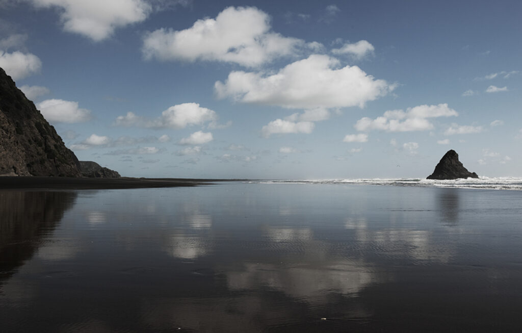 Beach with Black sand , solitory rock in surf with clouds annd blue sky reflected