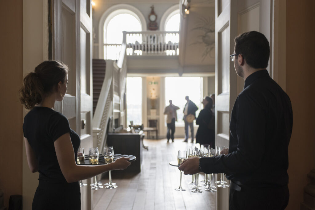 Waiters holding drinks for guests in background of out of focus Ragey Hall stairs